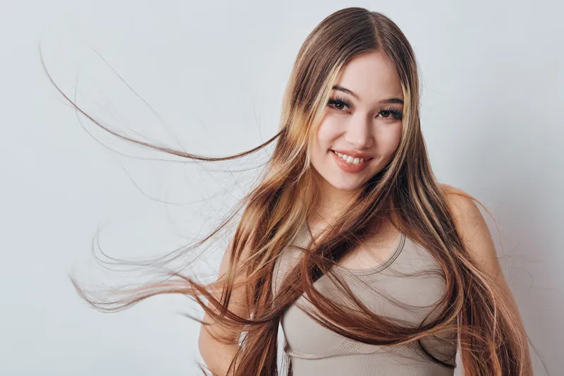 Young woman with long hair smiling at camera wearing casual tank top with light background. Beauty portrait with natural makeup and flowing hair for lifestyle concept.