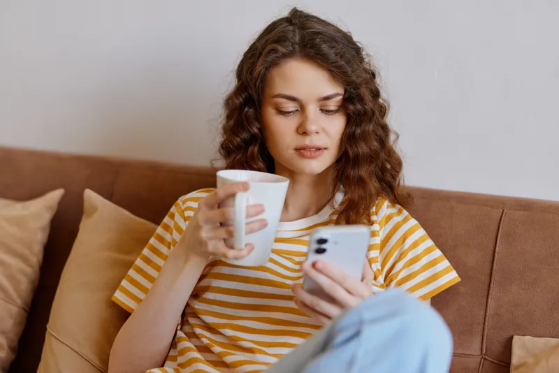 Casual woman at home sits on a couch, checks her smartphone while holding a mug, wearing a yellow striped shirt, relaxed mood, everyday life in a cozy living room.