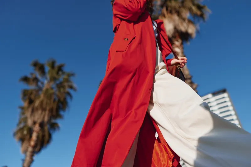 A bold outdoor fashion shot features a red coat and flowing dress in a street setting, wind lifting fabric under a bright blue sky, showcasing confident streetwear style.