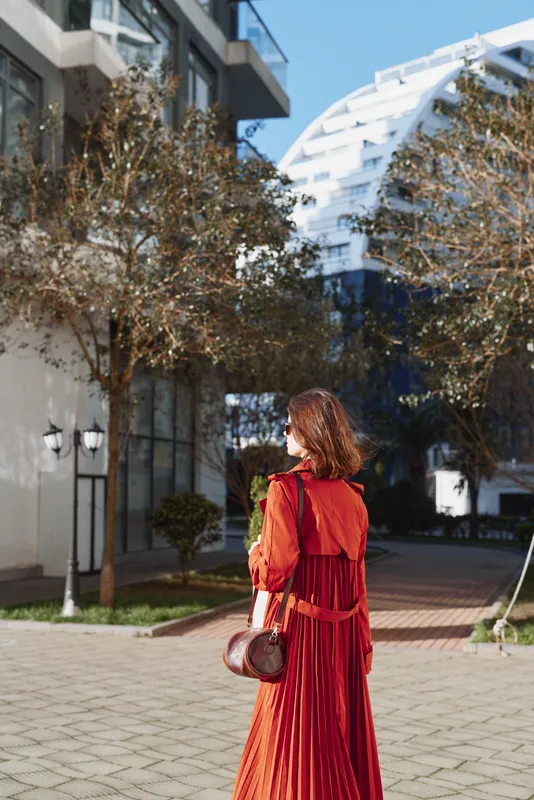 A woman in a flowing red dress walks along a sunny city street, looking over her shoulder as modern glass buildings frame the scene, capturing urban elegance and confident style.