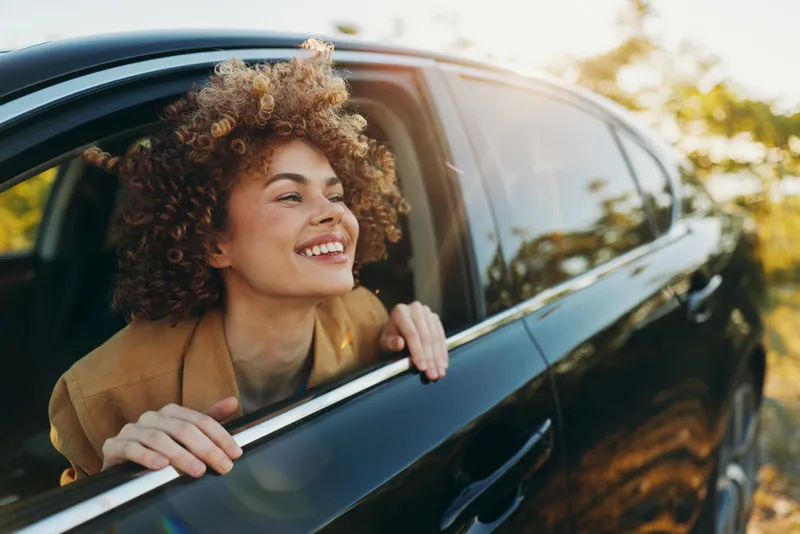 A young woman with curly hair leans out of a car window during a sunny drive, smiling as the retro color palette gives a carefree, cinematic mood.