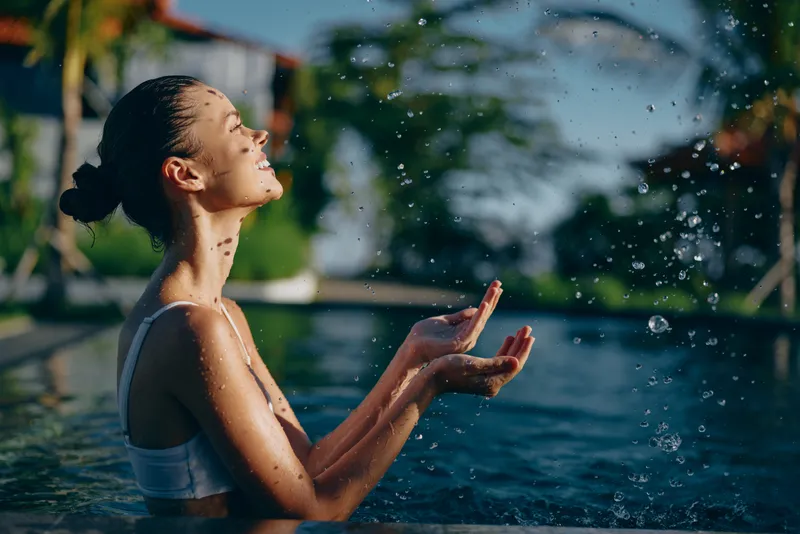 A woman enjoys water in a tropical outdoor pool under bright sunlight, cupping hands as droplets rise, capturing a carefree leisure moment by the pool.