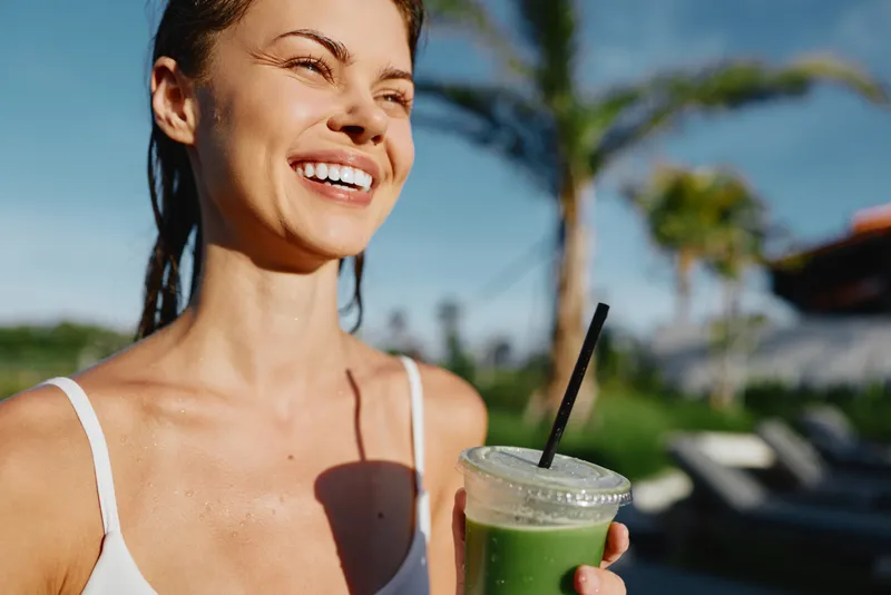 Young woman with short hair enjoys a refreshing green smoothie outdoors on a sunny day, smiling joyfully with palm trees in the blurred background.
