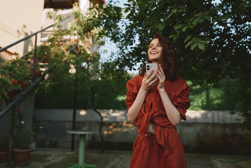 Bright outdoor lifestyle portrait of a smiling woman in a casual orange dress, holding a smartphone, amid greenery in a garden courtyard with a relaxed, joyful vibe