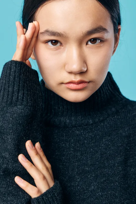 Portrait of young woman with dark hair wearing black turtleneck sweater against light blue background with thoughtful expression and hand touching face in studio.