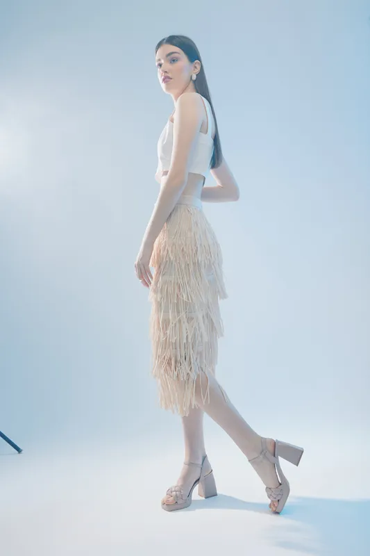 Elegant woman in a stylish fringed skirt and white top poses in a bright studio. The image focuses on fashion, beauty, and confident female style with soft lighting.