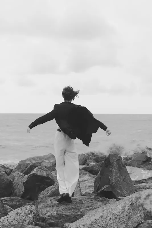A lone figure stands on rugged rocks by the sea, wind sweeping the coat as waves crash, creating a dramatic monochrome landscape of freedom