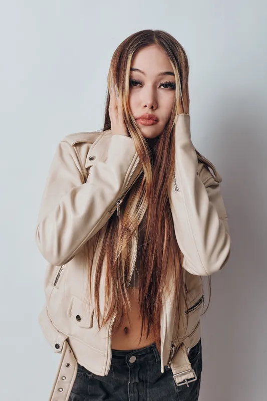 Young woman with long hair wearing beige jacket and black pants posing with hands on face against white background, portrait of fashion and casual style model.