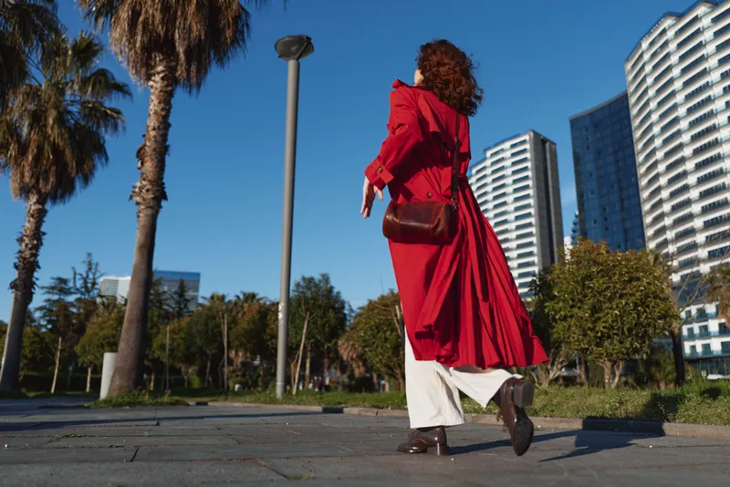 Woman in red coat walks on city street with modern buildings and palm trees around, carrying brown leather bag, sunny day with clear blue sky.