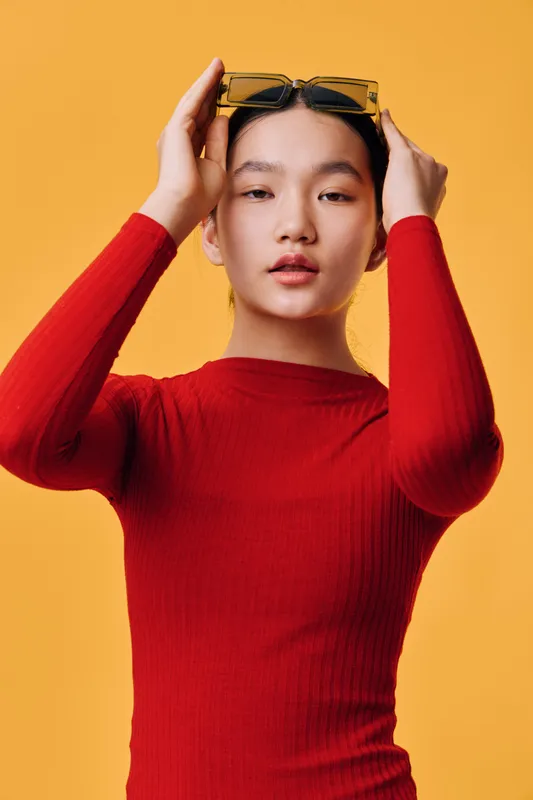 Young woman with long sleeve red shirt adjusting yellow sunglasses on head. Portrait with smooth skin and relaxed facial expression on orange background in studio light.
