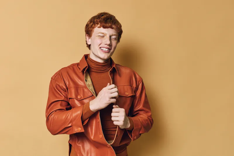 Young man with red hair wearing brown leather jacket and turtleneck smiling with eyes closed on beige background in studio portrait.