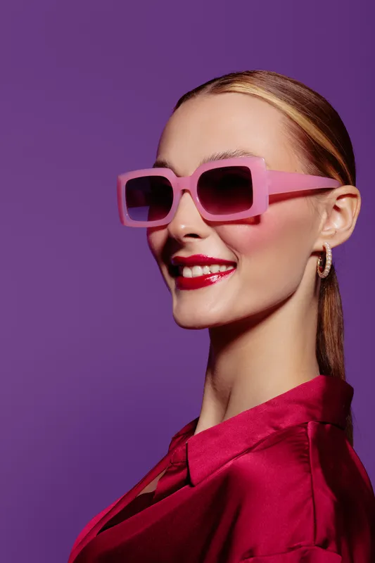 Smiling woman with pink sunglasses and red lipstick poses on purple background wearing shiny red blouse. Concept of fashion, beauty, style, and confident female portrait.