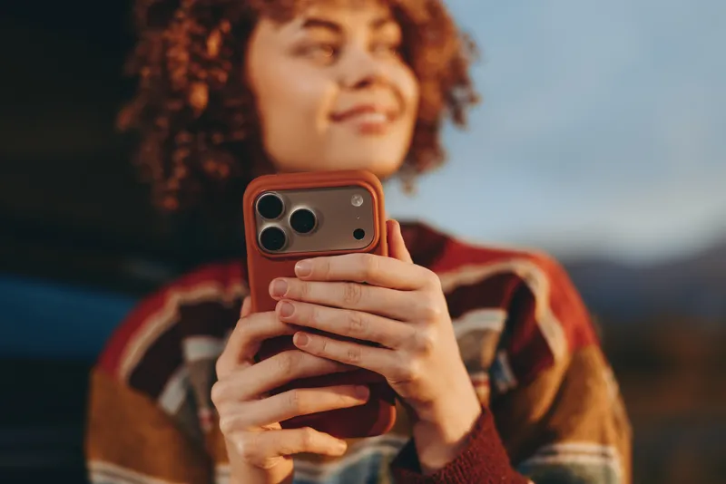 Woman lifestyle portrait outdoors, curly hair, holding smartphone and phone for selfie, relaxed smile, casual jacket, soft evening light, blurred background, modern technology