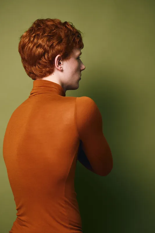 Young person with red hair wearing orange turtleneck sweater standing sideways on green background in studio. Profile, casual clothing and portrait of thoughtful individual.