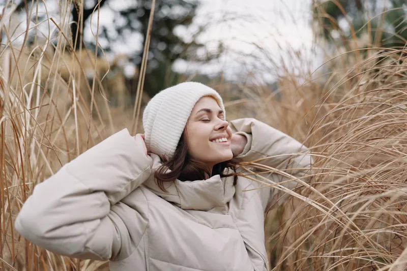 Woman in a warm beige coat and knit hat enjoying outdoor winter scenery among tall grass, relaxed smile, soft natural light, candid portrait capturing comfort and seasonal calm.