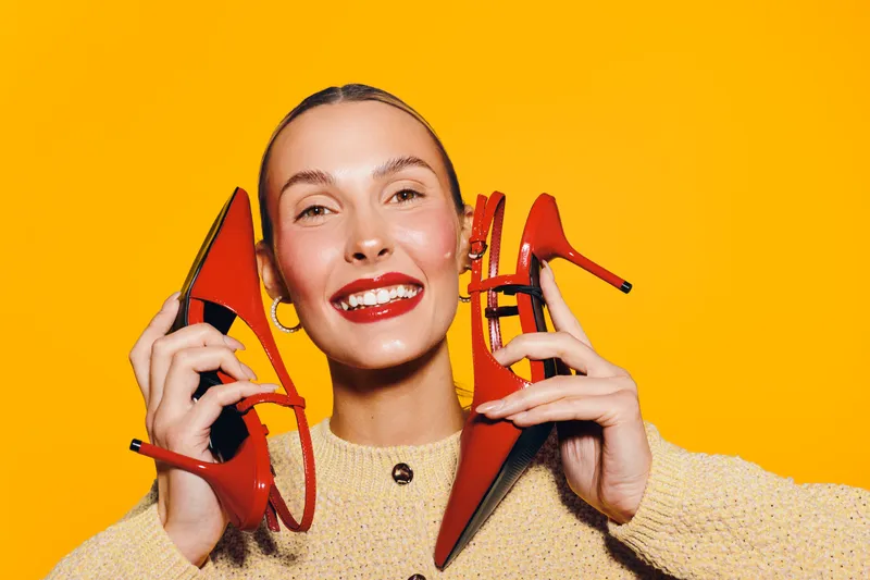 Smiling woman with red lipstick holds two red high heel shoes near her face on bright yellow background. Concept of fashion, style, beauty, and happiness with vibrant colors.