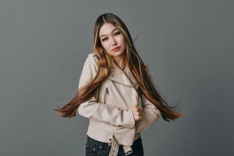 Young woman with long hair wearing light jacket posing on gray background, looking at camera with natural expression in studio portrait, casual fashion style.