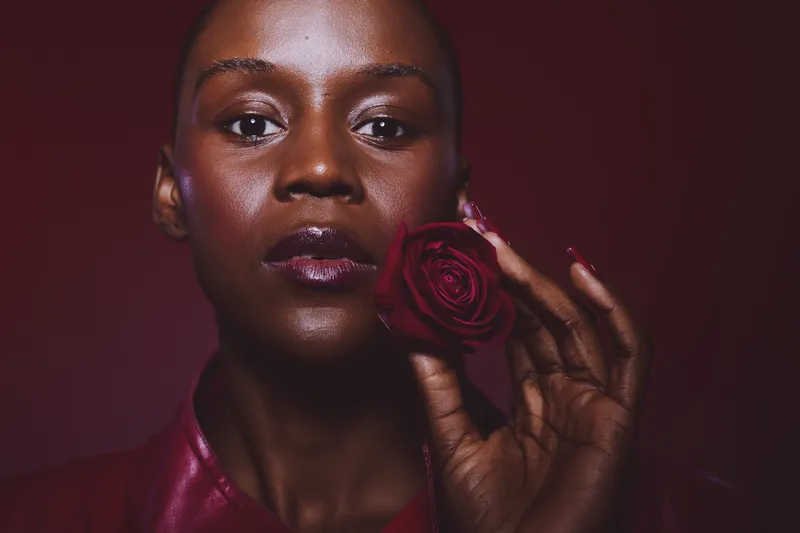 Close-up of a dark skinned African woman holding a red rose near her face. The portrait features glossy lips and smooth skin with deep red background and soft lighting. Beauty concept.
