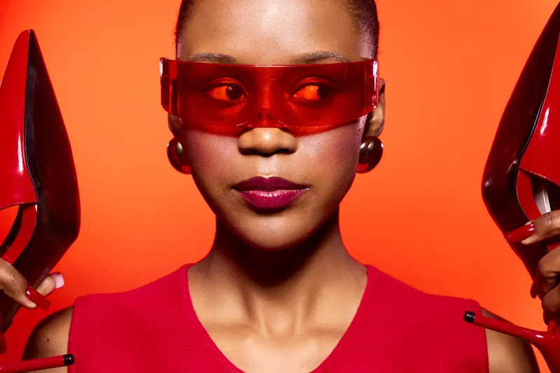 Close-up of a confident black woman with dark skin wearing red sunglasses and a red dress holding red high heels against an orange background. Concept of fashion and bold style.