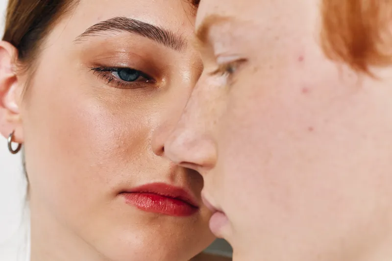 Closeup portrait of two young women with natural skin, focusing on facial details including acne and makeup, showing diversity and skin texture in soft lighting.