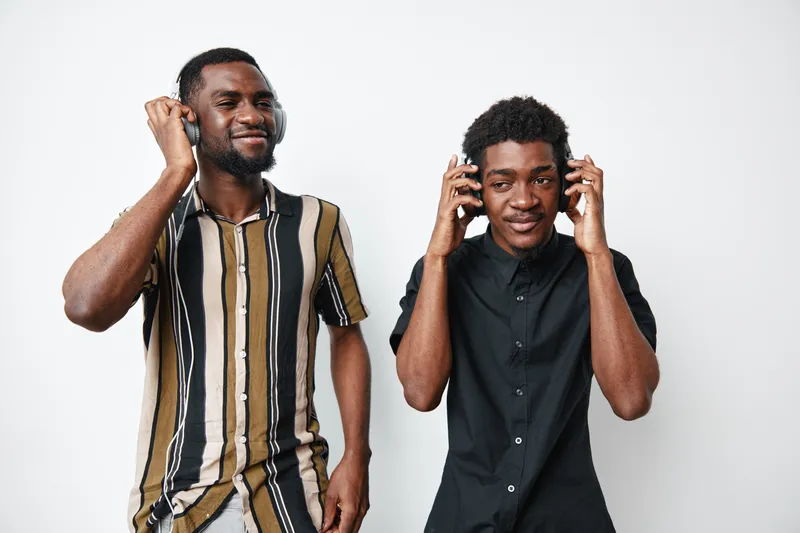 Two young black men with dark skin listening to music on headphones standing against white background. Casual style, smiling and enjoying sound together in studio.
