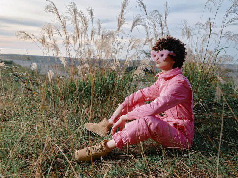 Woman in a pink outfit wearing a decorative mask sits in tall grass outdoors, enjoying a calm moment in an open landscape with boots and wild plants.