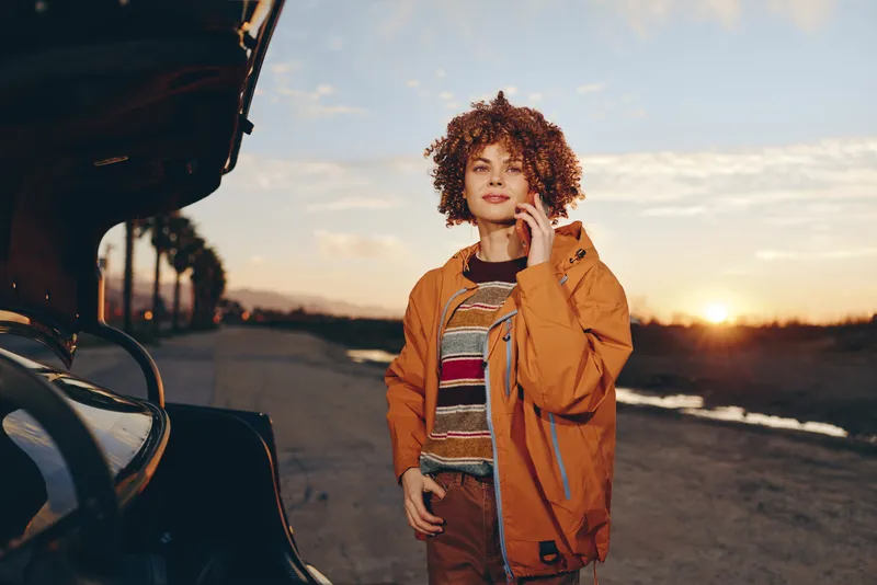 Woman with curly hair posing near a car at sunset, outdoor lifestyle scene on a road, wearing an orange jacket, soft evening light, travel feeling relaxed mood