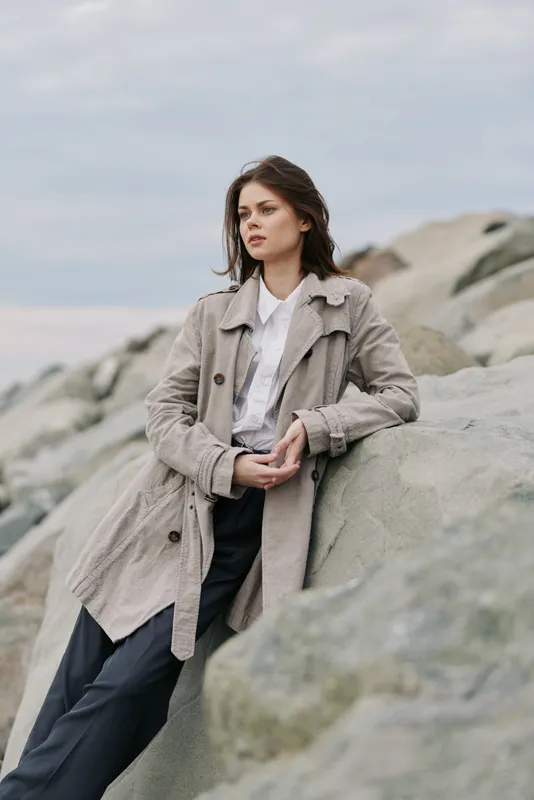 Young woman in casual coat leans on large rocks outdoors with cloudy sky background. Natural portrait of confident model in relaxed pose near water and stone landscape.