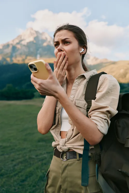 Young woman on a hike checks her phone in a sunlit valley, surprised by a message, while wearing a beige shirt and olive pants and carrying a backpack.