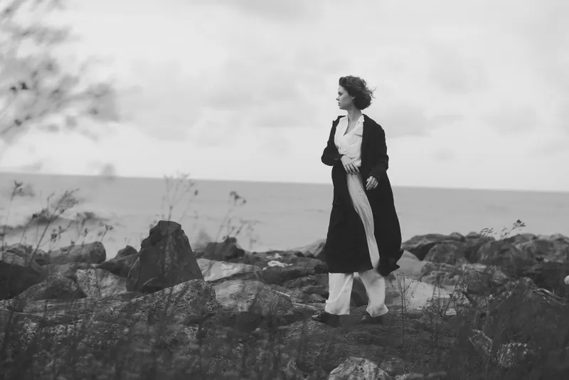 Woman on a rocky coast in a long coat walking along the shore, black and white scene with wind swept hair and calm sea horizon