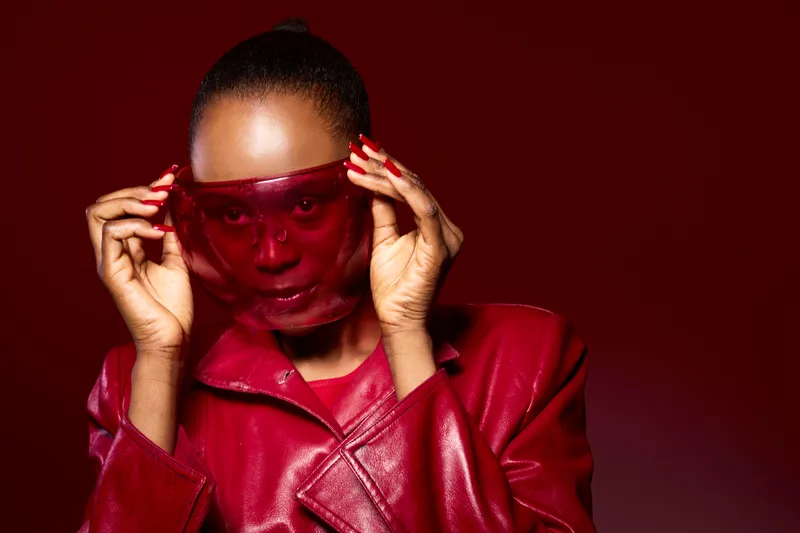 Portrait of a dark skinned African woman wearing a red leather jacket and red translucent mask. Studio shot with red background. Concept of fashion, style, and bold accessories.