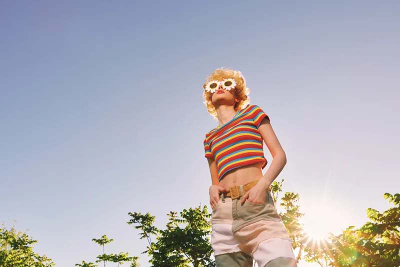 A young woman in a striped crop top and high waisted shorts poses outdoors with confident retro style under bright sun and blue sky.