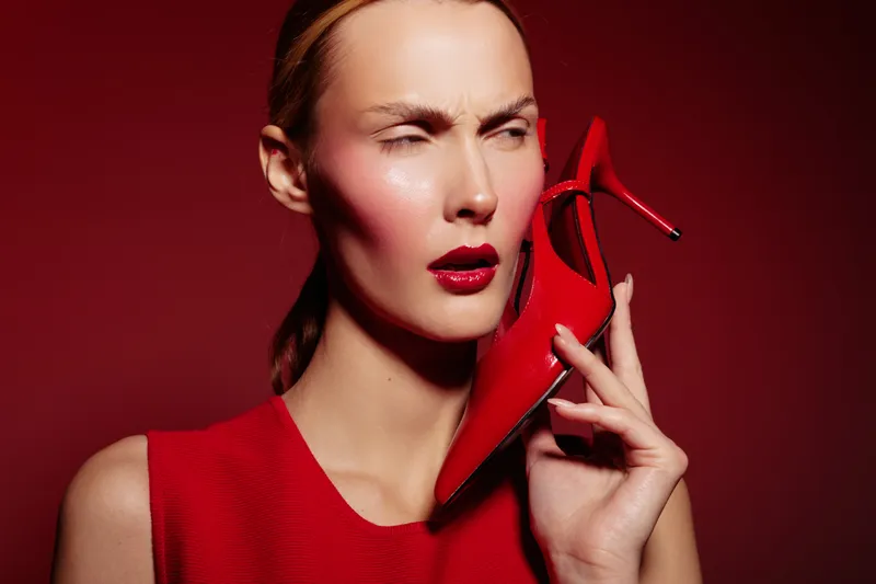 Woman in red dress holds red high heel shoe near face with confused expression on red background. Concept of fashion, style, and unusual accessories.