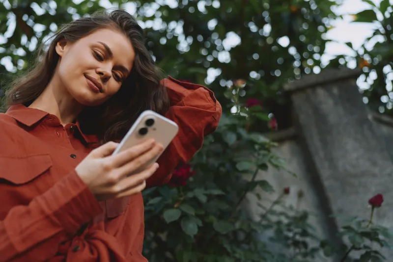 Young woman in a red shirt outdoors holds a phone, smiling softly as she browses, standing in a sunlit garden with blooming roses