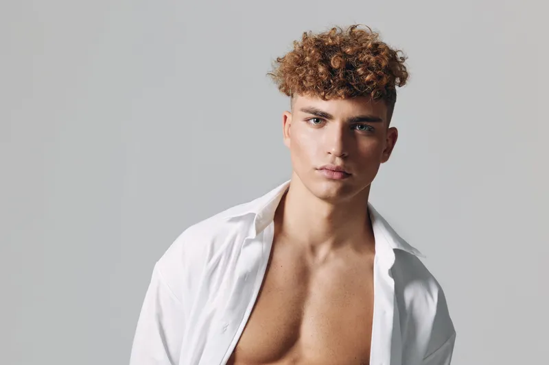 Young man with curly hair and open white shirt posing against gray background for fashion portrait and style concept with confident expression and casual look.