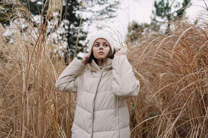 Woman in a beige puffer jacket stands among tall dry grasses in a chilly outdoor scene, conveying fashion and seasonal comfort in natural surroundings.