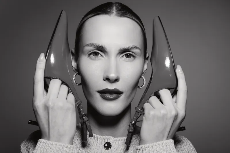 White woman with fair skin holds pair of high heel shoes near face looking at camera. Studio portrait in black and white with fashion and beauty concept.