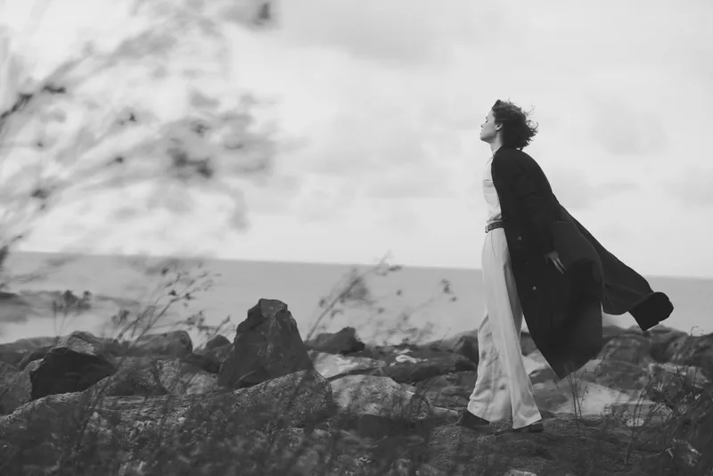 Windy coastal scene with a woman standing on rocks, cloak billowing, gaze toward the sea, solitude and exploration in a moody monochrome setting