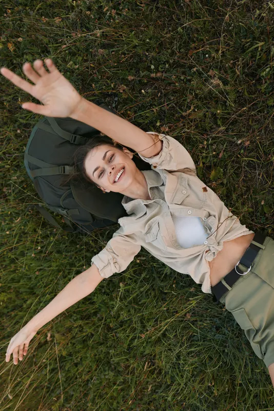 Young woman lies on green grass outdoors, smiling and relaxed with one arm stretched above her head, wearing a casual beige shirt and olive pants on a sunny day