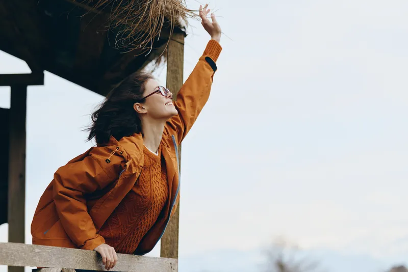 Woman lifestyle outdoors in warm daylight, orange jacket and knit sweater, leaning on wooden fence, one arm raised, hair lifted by wind, gazing at clear sky, relaxed mood today now