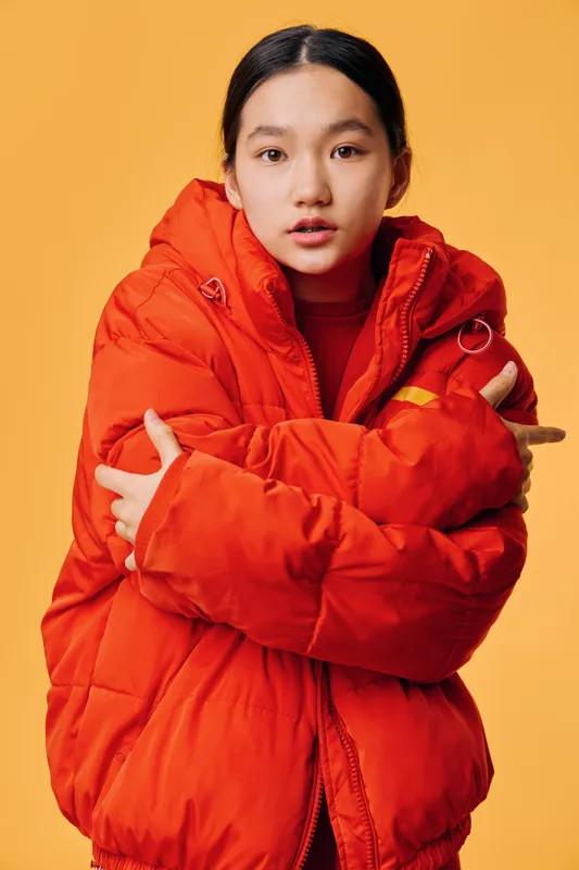 Young woman wearing bright orange puffer jacket stands against yellow background with arms crossed. Studio portrait of casual fashion, winter clothing and confident expression in warm style.