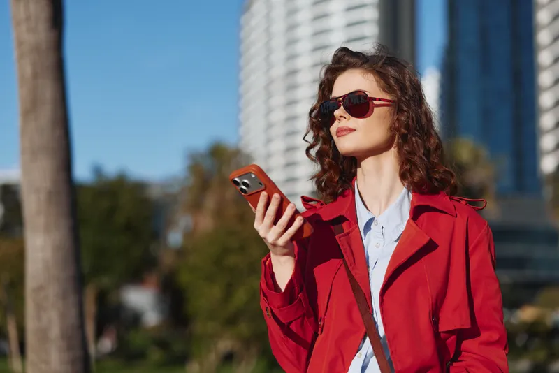 A stylish woman in a red coat and sunglasses stands outdoors, looking at her phone with a calm, confident expression against a bright urban backdrop.