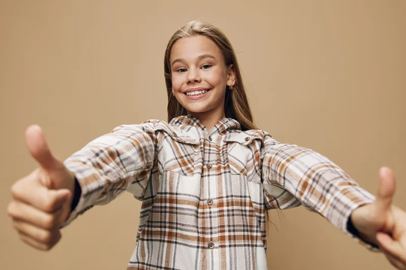 Girl smiling and showing thumbs up in plaid shirt with long hair standing on beige background. Happy child expressing positivity and confidence with cheerful facial expression.