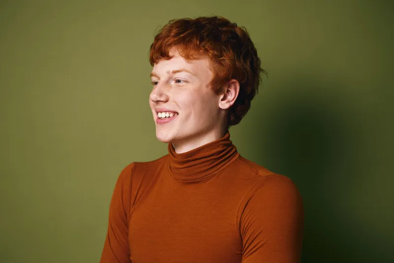 Young man with red hair wearing brown turtleneck smiling and looking away against green background in studio portrait with natural expression and casual style.