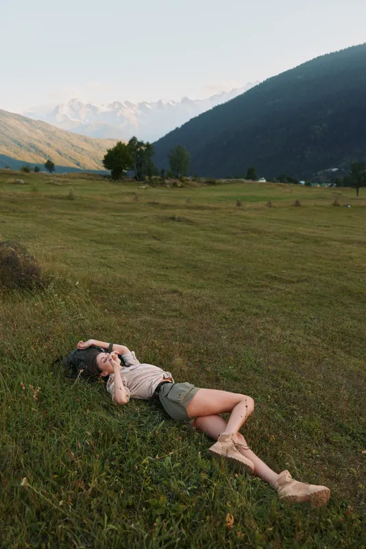 Relaxed woman lying in a green meadow with distant hills, enjoying nature and sunshine. Casual outfit highlights outdoor lifestyle and a peaceful, carefree moment and mood.