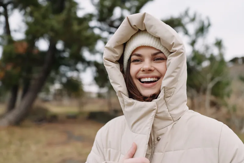 Smiling woman wearing a beige puffer jacket with a hood and knit beanie, outdoors in a cool natural setting, capturing a candid winter moment in nature