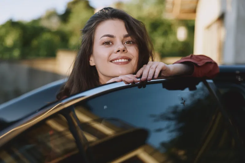 A young woman leans on a sleek car, smiling at the camera in bright daylight, conveying a relaxed, confident mood and outdoor lifestyle