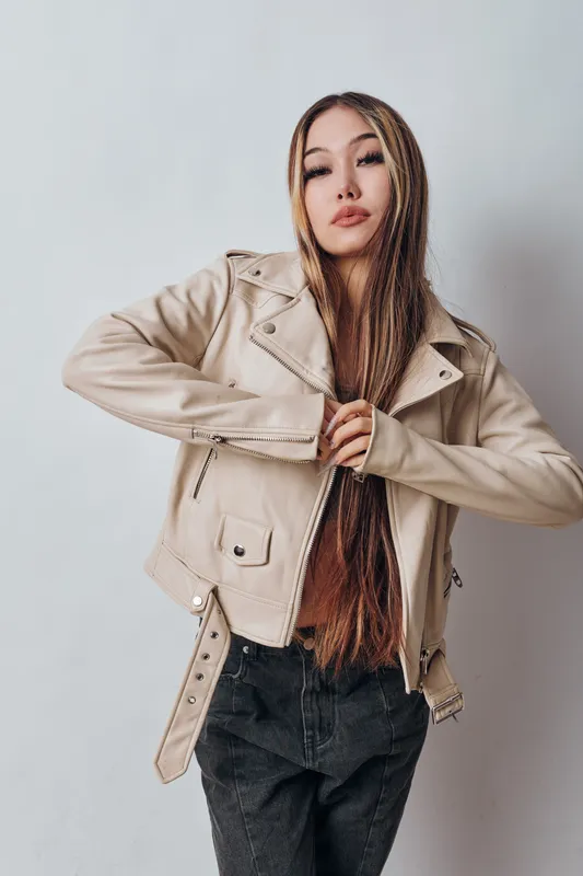 Young woman with long braided hair wearing beige leather jacket and black pants posing against plain light background with confident facial expression and stylish look.