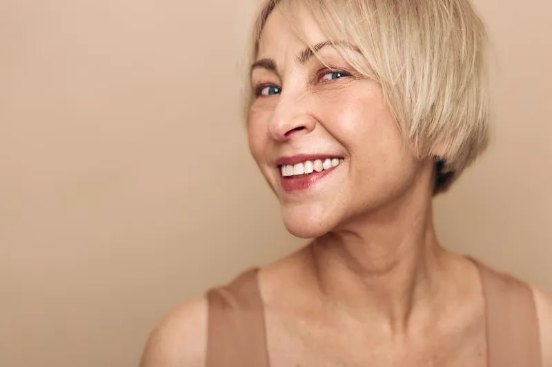 Smiling middle aged woman with short blonde hair looks confident and happy on beige background. Portrait of natural beauty, wellness, and positive emotion with soft lighting.
