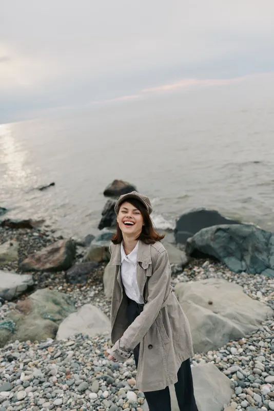 Young woman in coat and hat laughs happily on rocky beach near calm sea under cloudy sky. Casual autumn fashion and joyful mood in natural coastal landscape.
